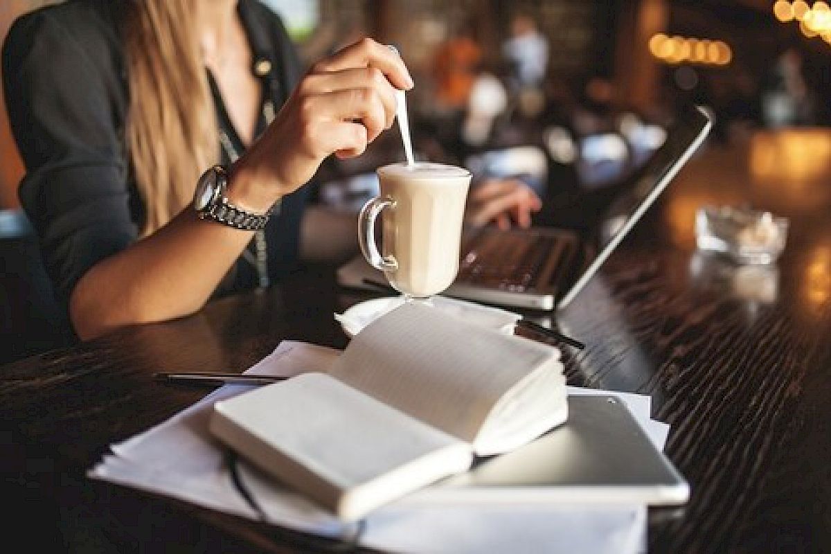 A person in a cafe is stirring a drink while using a laptop, with notebooks and a smartphone on the table.